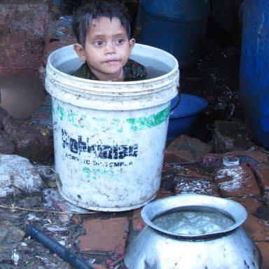 A young child sits in a white plastic bucket filled with water, surrounded by various containers and items on a wet, muddy surface. The child's expression is calm as they look off into the distance. The bucket has faded text and a green stripe.