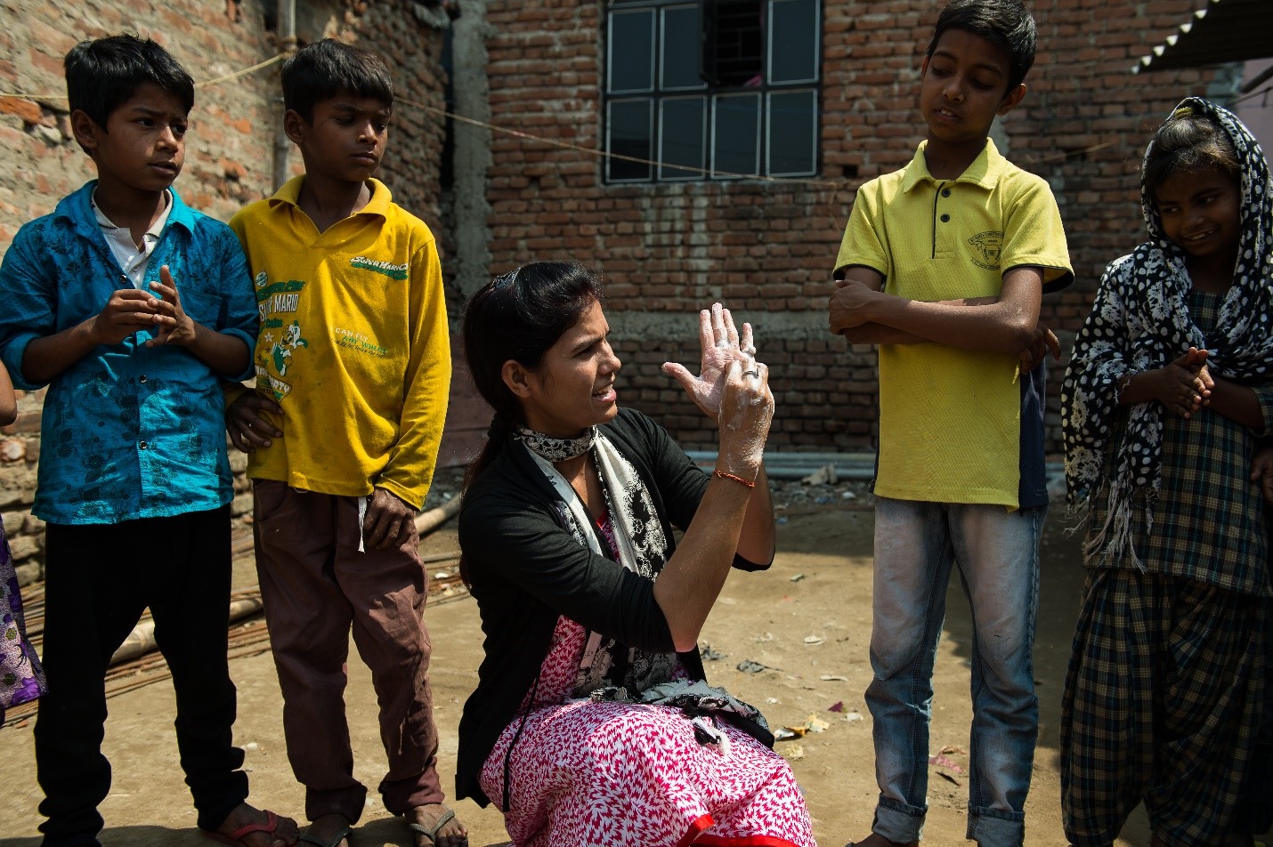 A woman squats on the ground, using sign language to communicate with a group of children gathered around her. The children look attentively at her gestures. They're standing in an open area, with brick buildings in the background. The woman is wearing a pink scarf.