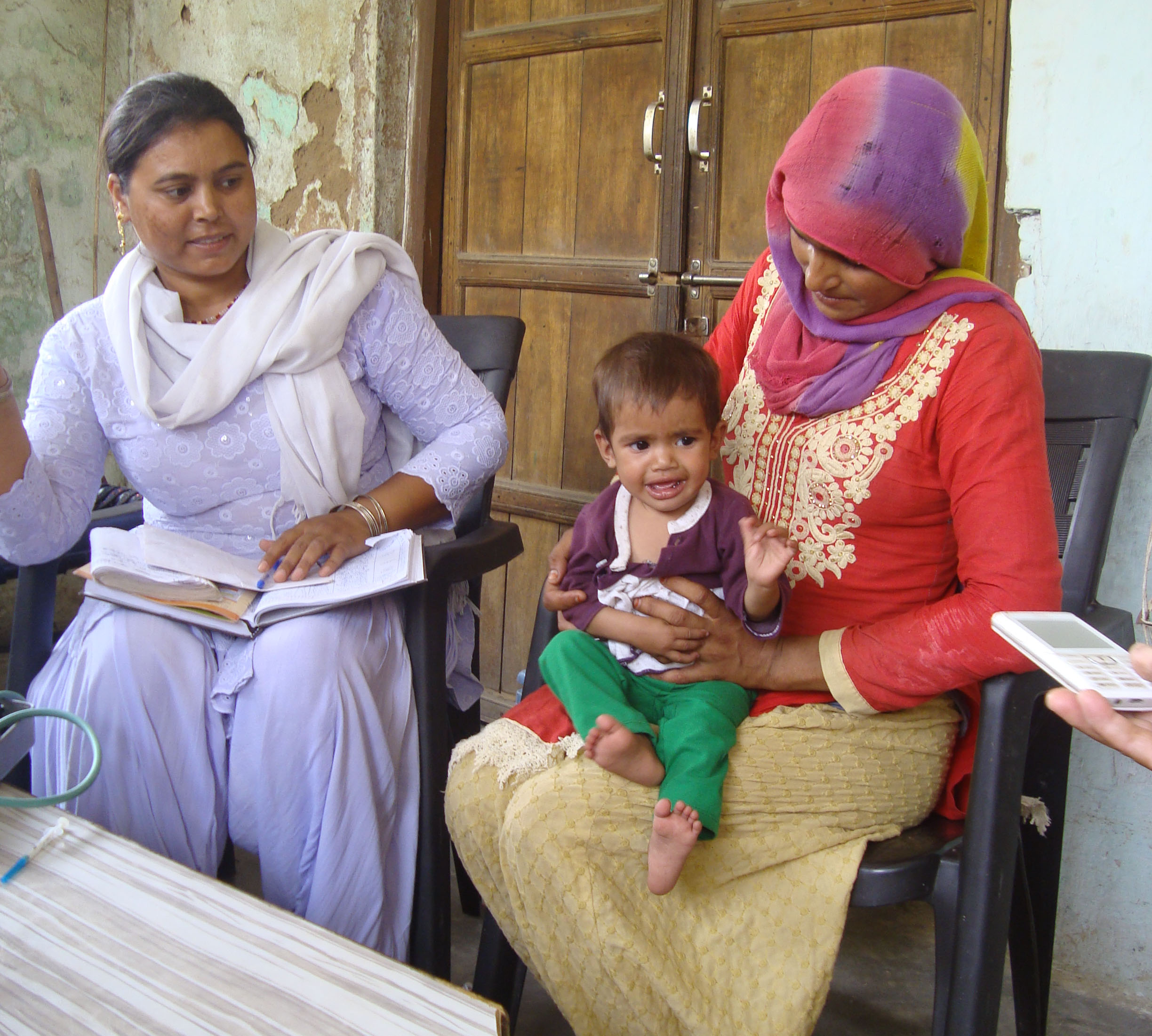 Two women sit in a room, one holding a child and the other holding a book. The woman on the left wears a white outfit, while the woman on the right, wearing a red and yellow outfit with a purple headscarf, holds a smiling toddler on her lap.