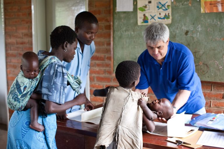 A healthcare worker in a blue shirt assists a child lying on a table while two adults and a child observe. Another child is carried on the back of an adult. The setting is a clinic with a brick wall and chalkboard in the background.