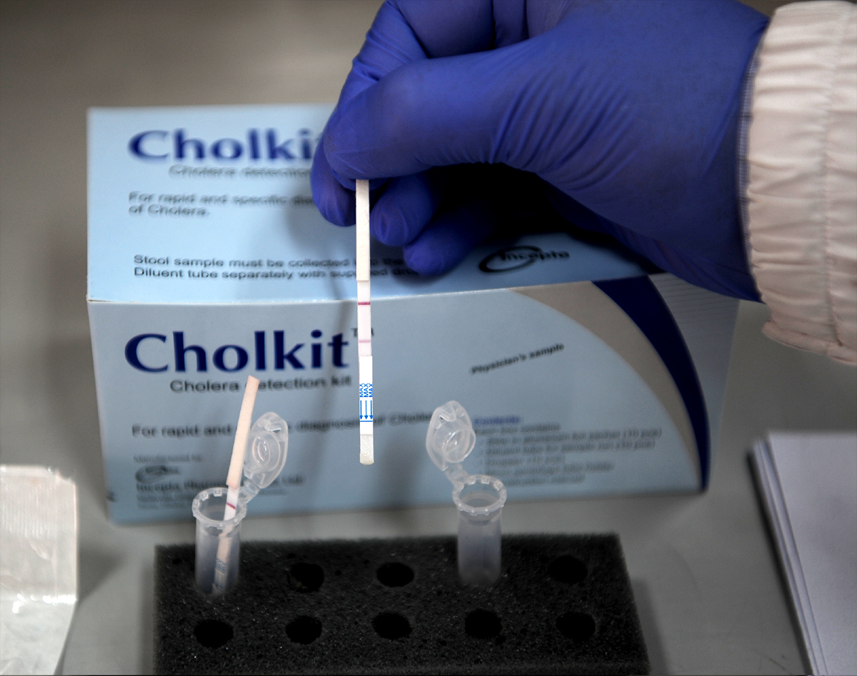 A gloved hand holds a Cholkit test strip above a testing station. Two small vials are set in a foam holder. Behind, the Cholkit packaging is visible, indicating that it's a cholera detection kit.