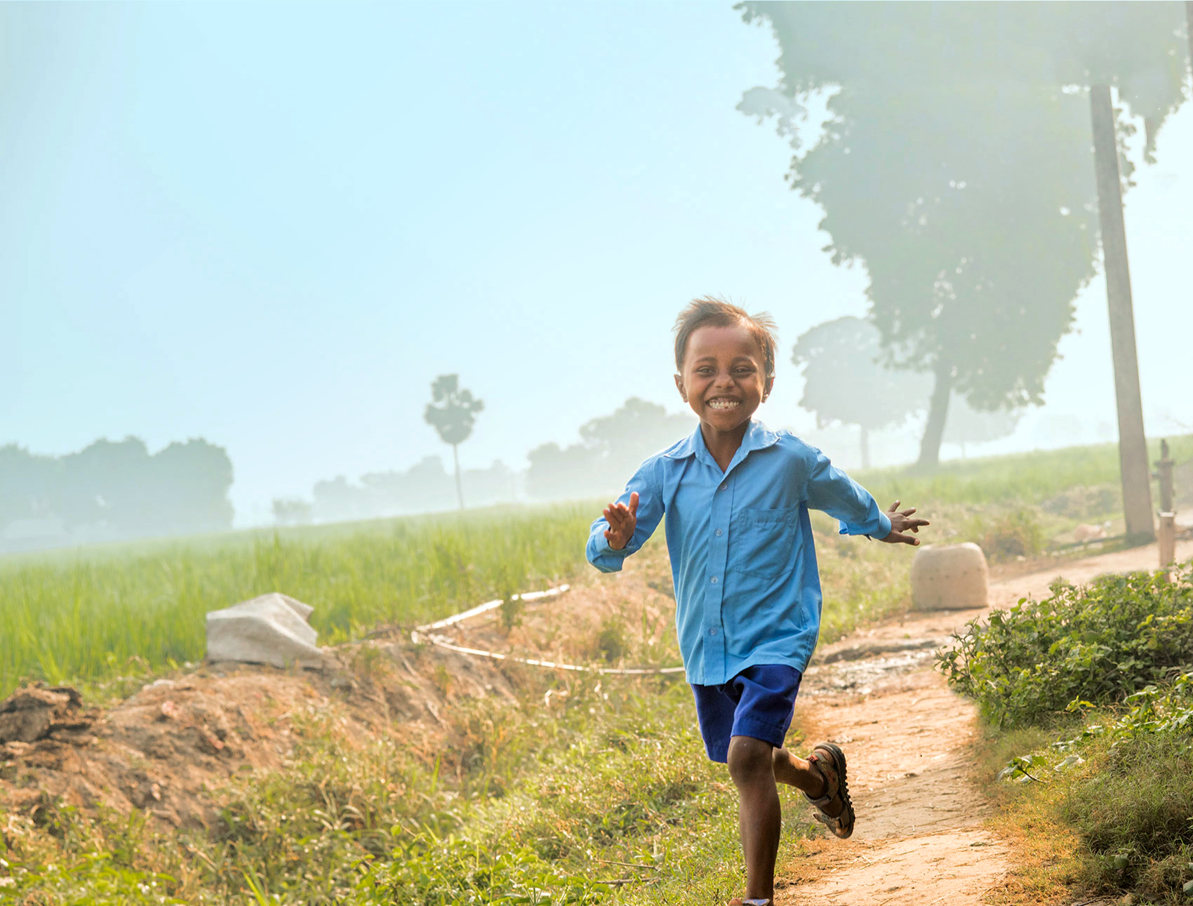 A joyful young boy in a blue shirt and navy shorts runs along a dirt path through a lush green field. The background is filled with misty trees and a hazy sky, creating a serene rural scene.