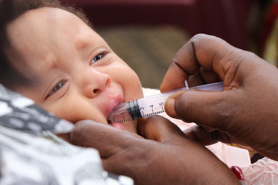 A close-up of a baby with light skin and blue eyes being fed liquid medicine or nutrients through an oral syringe by an adult's hand. The baby appears to be lying down and looks slightly uncomfortable.