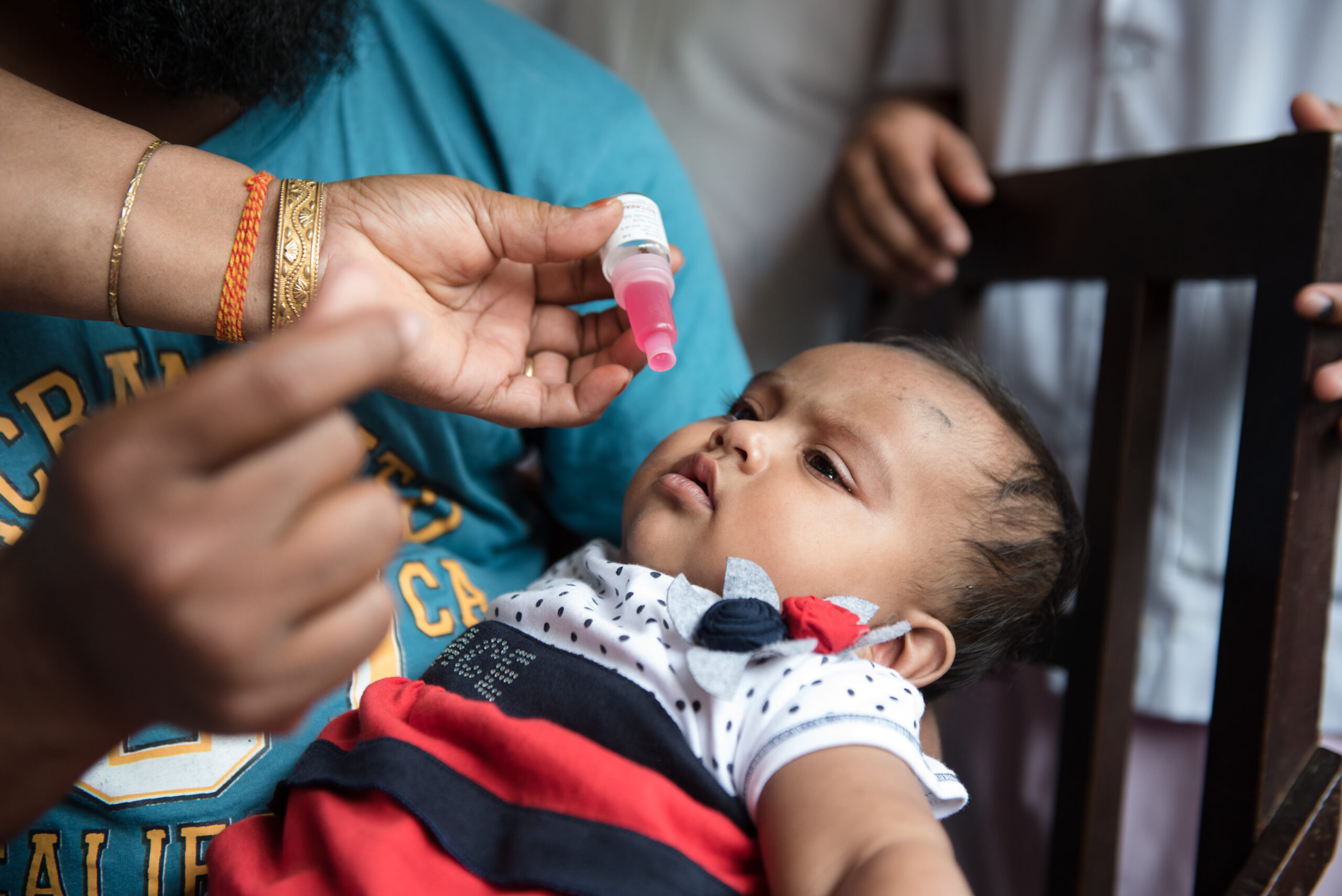 A baby wearing a striped outfit lies in an adult's arms, looking up at a small bottle held by another person, who is about to administer liquid drops. The adult's hand is adorned with bangles, and other people are present in the background.