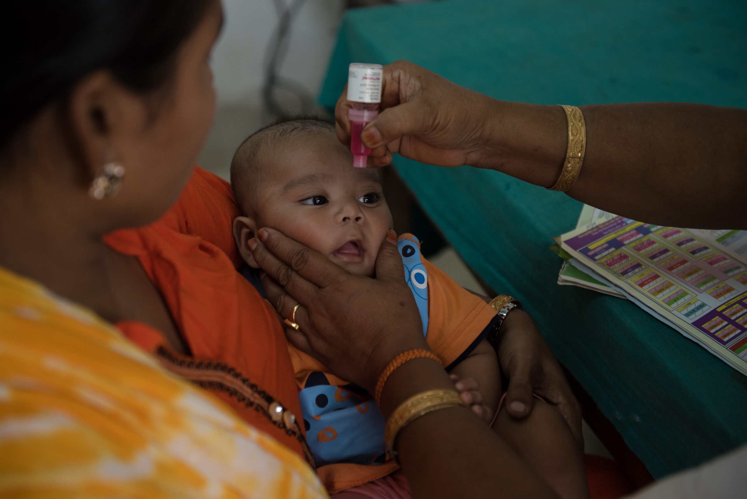 An adult holds a baby in their lap while another adult gives the baby a dropper dose of liquid medicine. The baby, wearing an orange outfit, looks up, and both adults’ hands are visible, with one supporting the baby's head and the other administering the medicine.