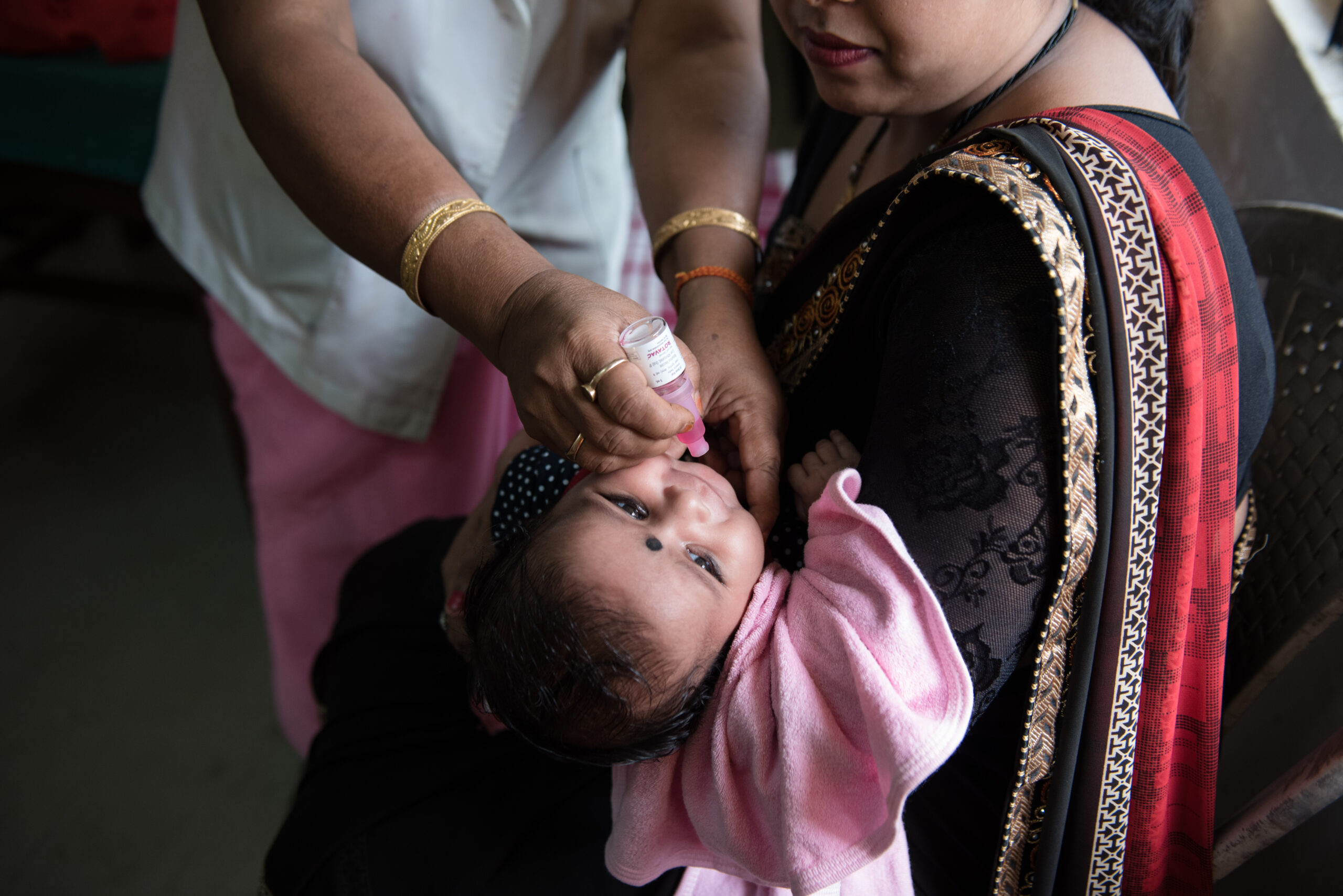 A woman dressed in a black saree with red and gold details holds a baby wrapped in a pink blanket. Another person administers an oral vaccine to the baby from a dropper.