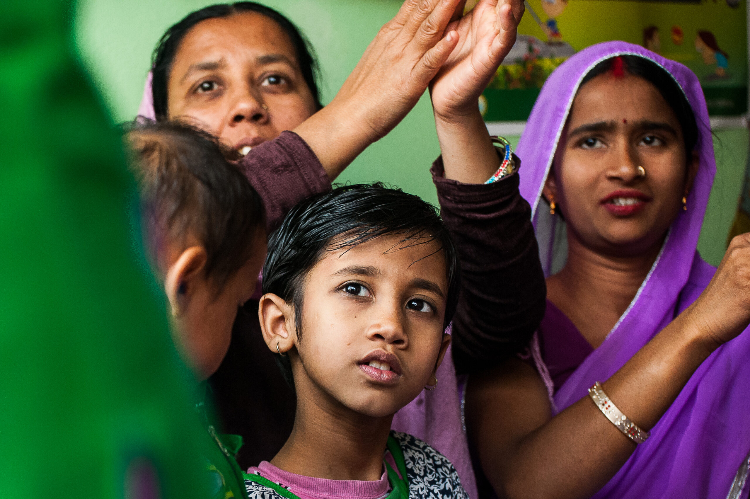 A young girl in a green shirt stands amidst adults raising their hands, including one woman in a purple sari. They appear to be in a lively indoor setting, possibly participating in an activity or event.