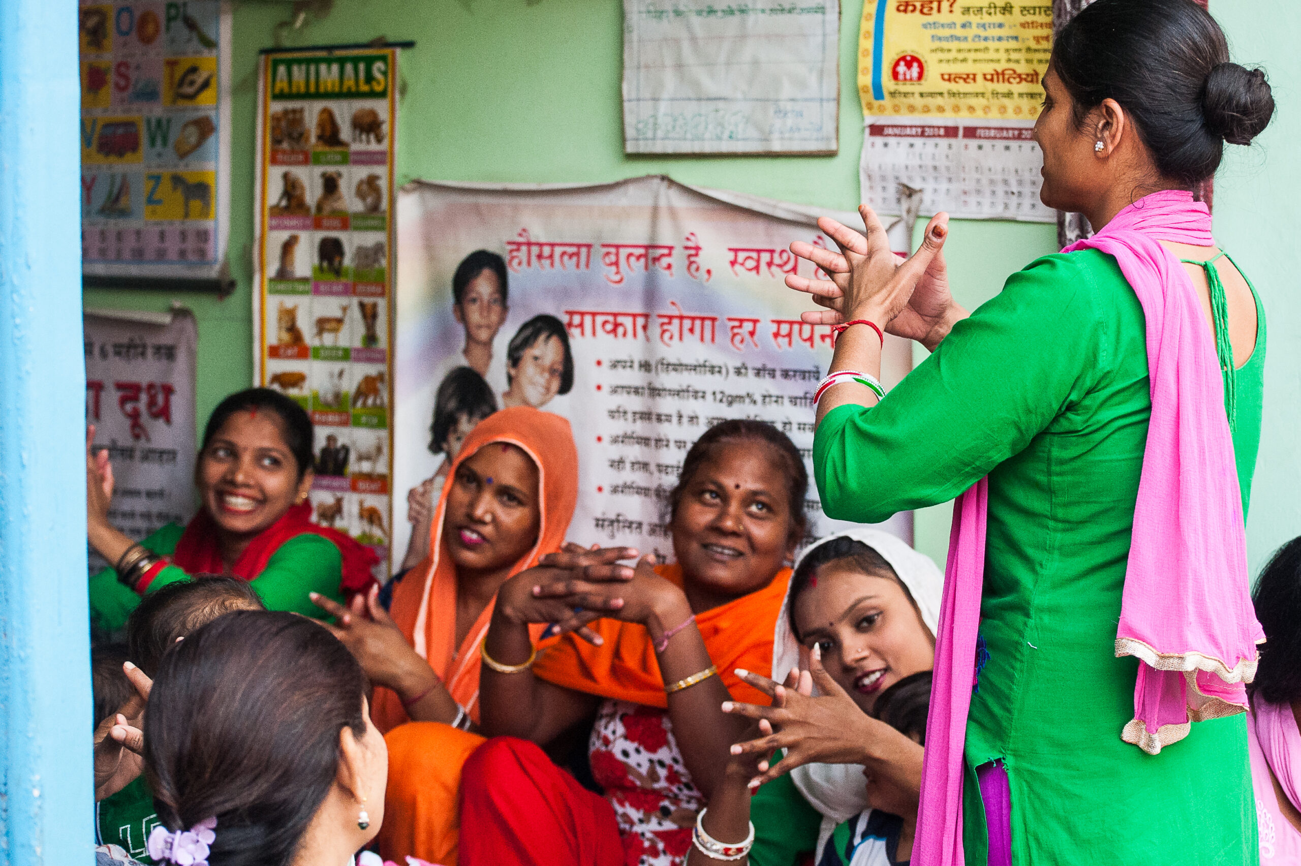 A group of women, clad in vibrant traditional attire, sit in a room attentively watching a standing female instructor. Posters with educational content adorn the walls, creating a lively and engaging learning environment.