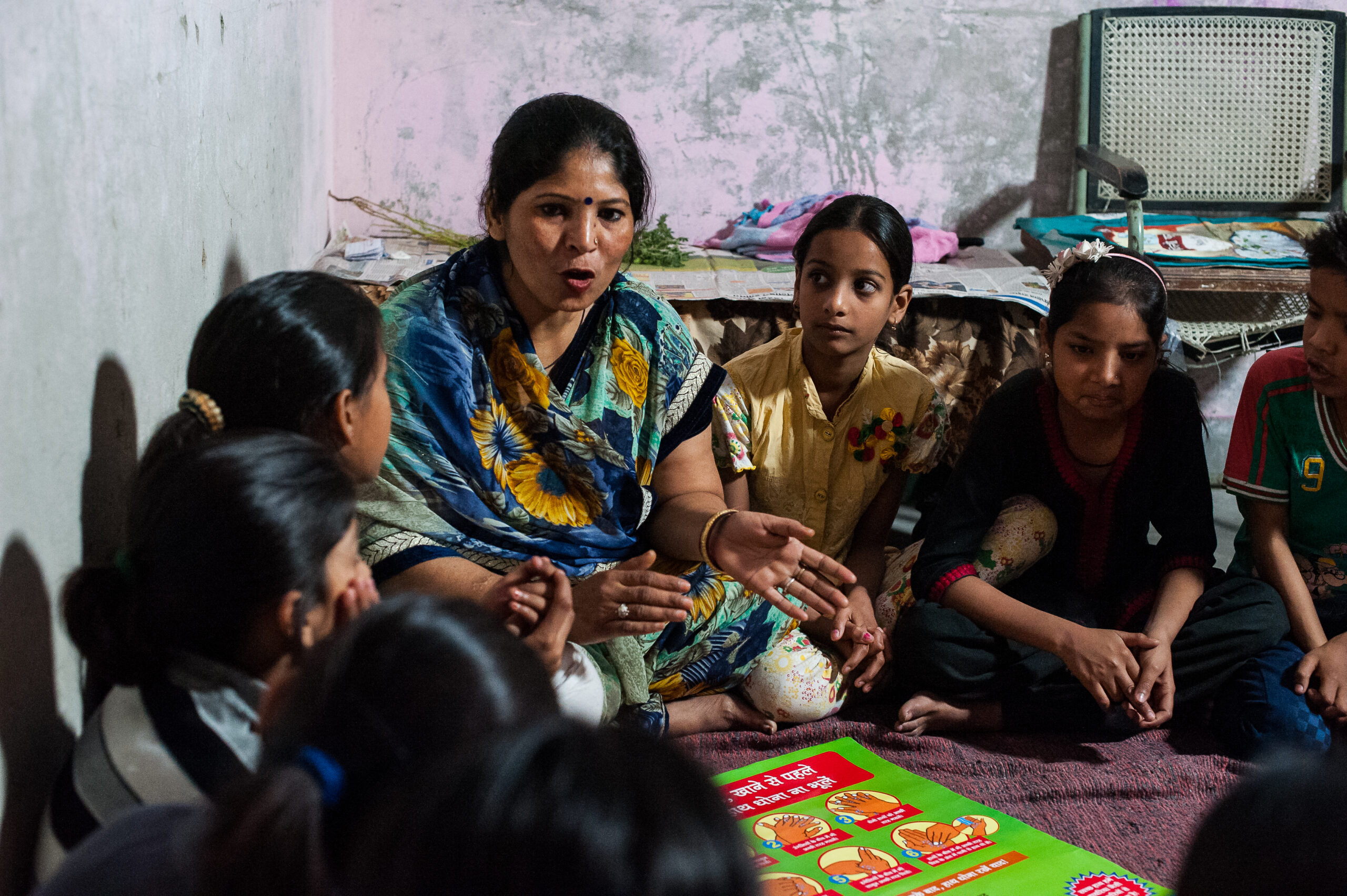 A woman is seated on the floor, surrounded by children, engaging them in a discussion. They are focused on a colorful educational poster. The setting appears to be a modest room with sparse furnishings.