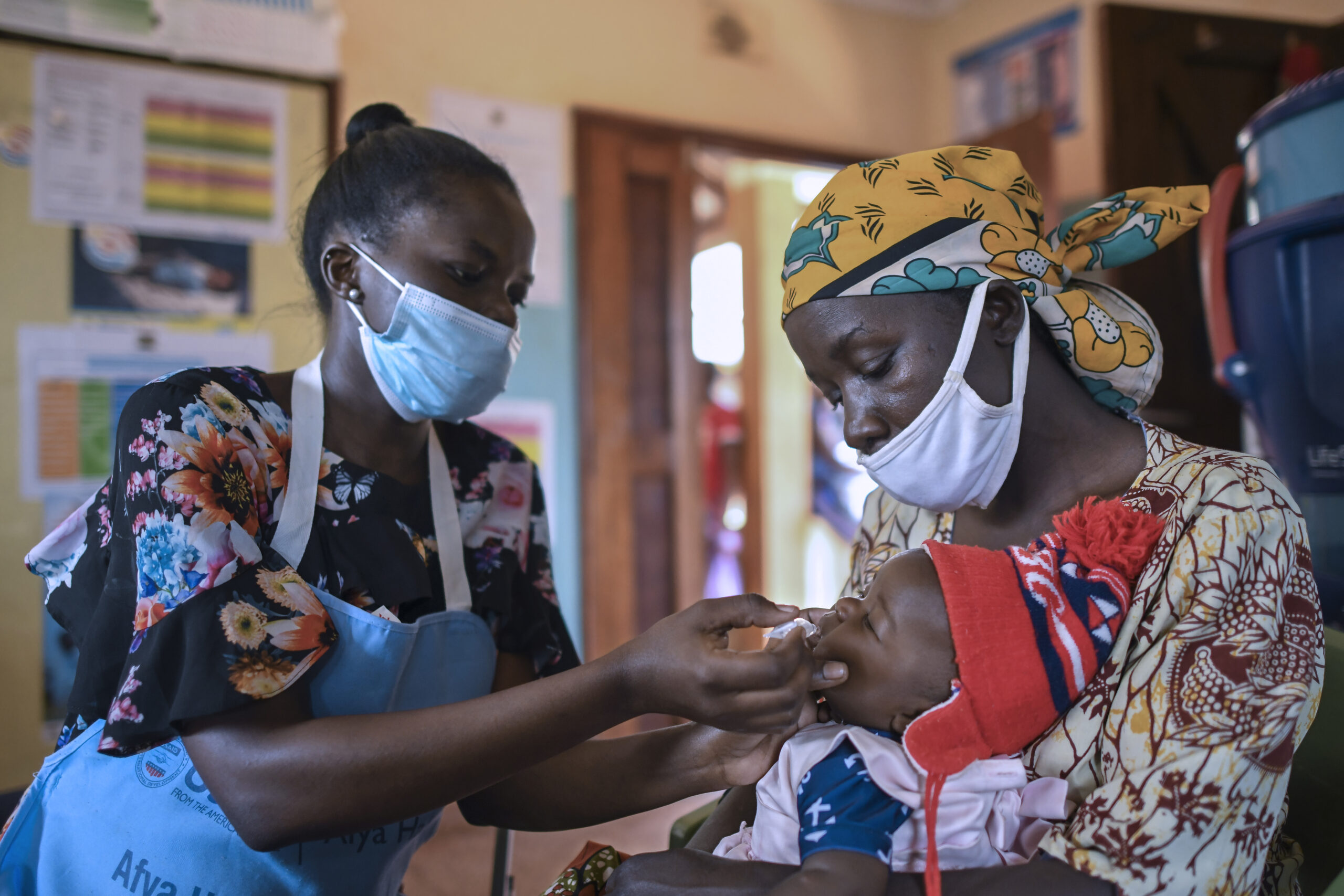 A woman is holding a child while another woman, both wearing face masks, administers an oral vaccine to the child. The scene appears to be in a healthcare facility with posters and charts on the walls in the background.