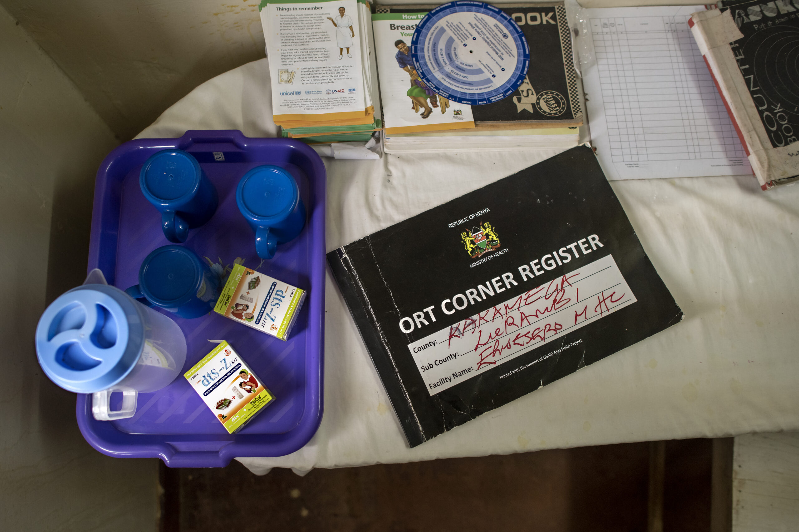 A medical register labeled "ORT Corner Register" lies on a white surface alongside a purple tray with blue cups, medication boxes, and a white bottle. Various notebooks and papers are partially visible nearby.