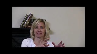 A woman with blonde hair is seated indoors in front of a light-colored wall. She is wearing a white top and gestures with her hands. Behind her on a dark shelf are several stacked books.