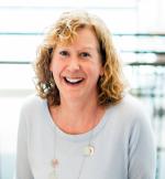 A woman with curly blonde hair and a big smile is wearing a light-colored top. She is sitting in front of a blurry background with horizontal lines.