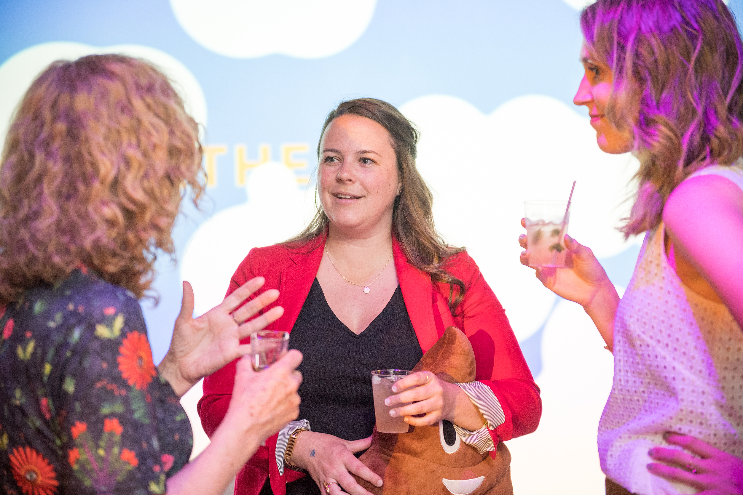 Three women are conversing and smiling while standing in a room with a playful blue, cloud-themed background. The woman in the center, dressed in a red blazer, holds a plush toy resembling a smiling poop emoji. All three have drinks in their hands.