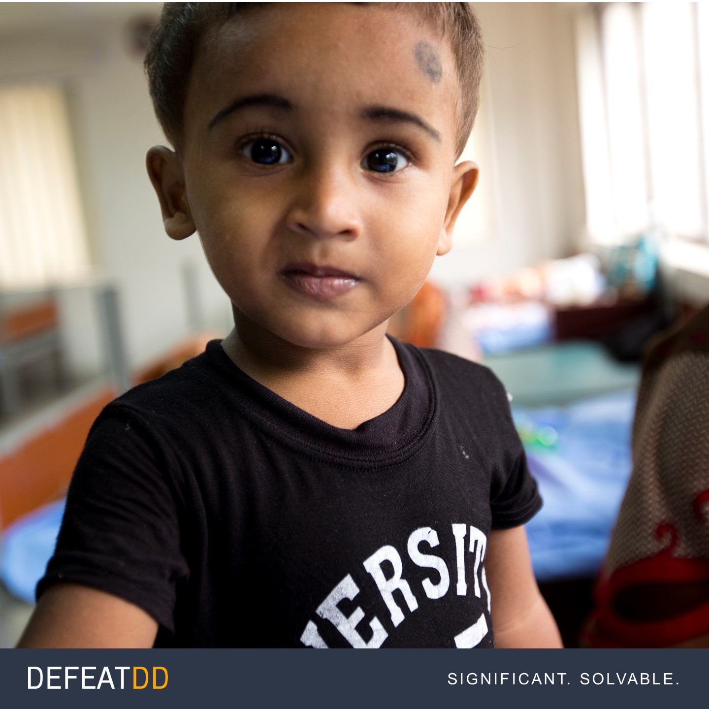 A young child stands indoors, wearing a black T-shirt with white text, and has a curious expression on their face. The background is slightly blurred, showing a room with benches and items. The image has the text "DEFEATDD: Significant. Solvable." at the bottom.