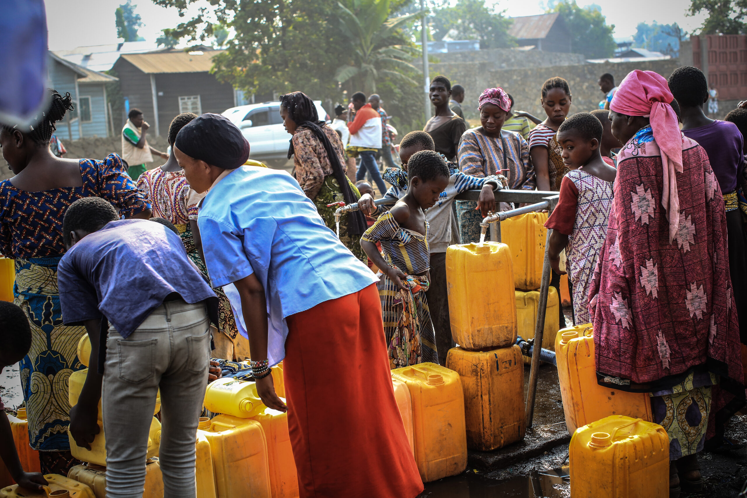 A large group of people, including children and adults, gather outdoors around many yellow plastic water containers. They are filling the containers with water, engaging in conversation, and working together. The setting appears to be a community water source in an urban area.