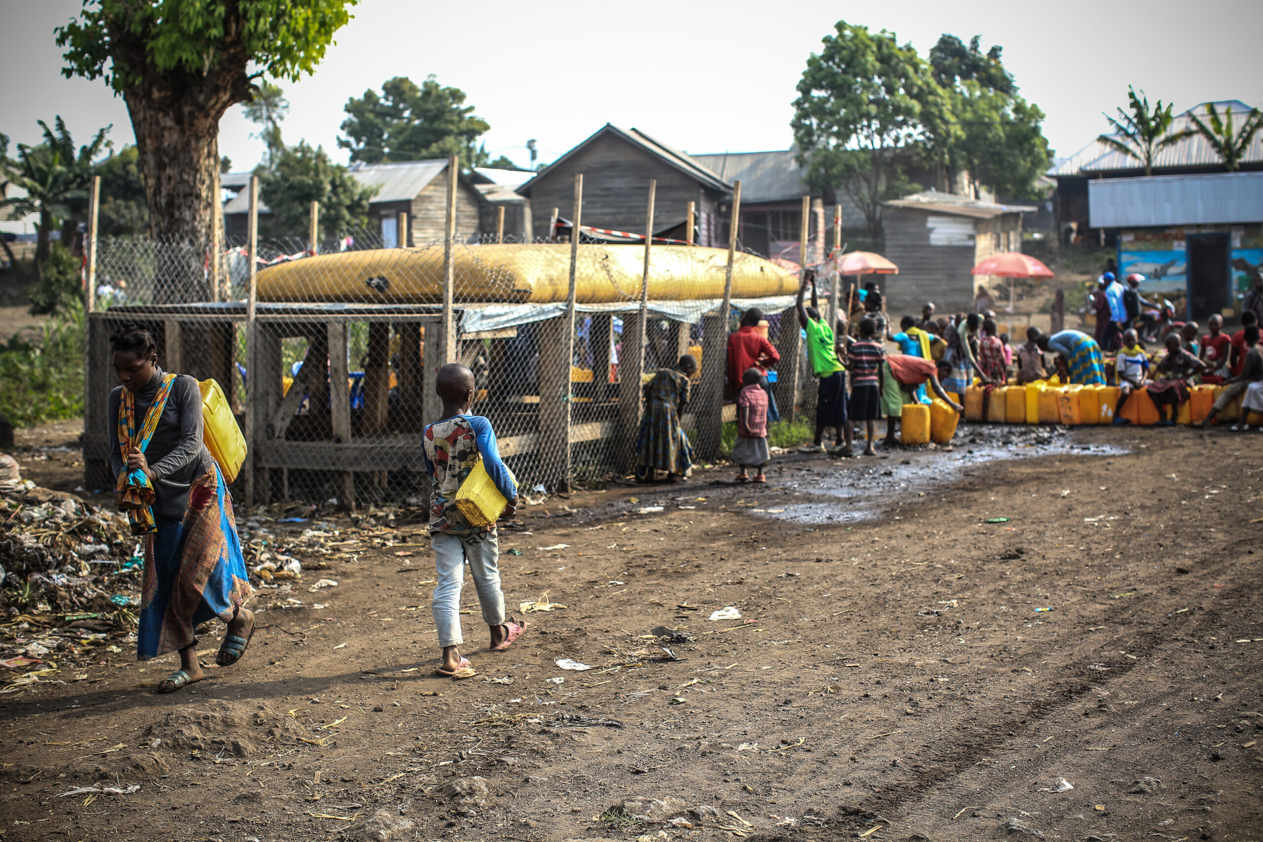 People gather near a water distribution point, carrying yellow jerry cans. The setting is a rural area with dirt ground, trees, and buildings in the background.