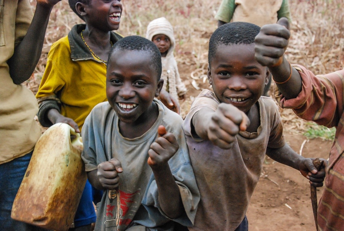 A group of smiling children outdoors, two in the foreground gesturing enthusiastically towards the camera. One child holds a plastic container. The background features more children and a dirt-covered, rural setting. The scene exudes joy and energy.