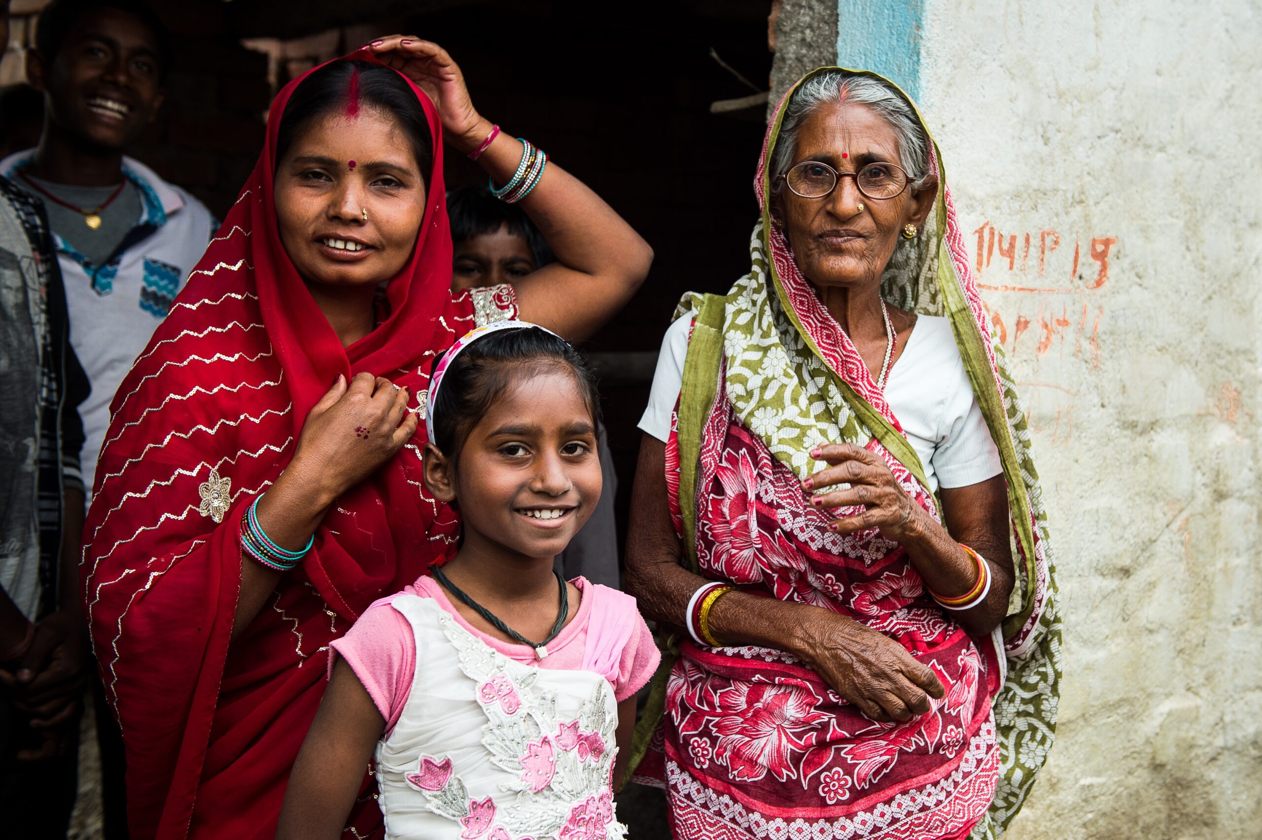 A young girl stands smiling in front of two women, one elderly and one middle-aged, all dressed in vibrant saris. They are positioned outside a building with the woman in red adjusting her scarf, and the woman in green wearing glasses.