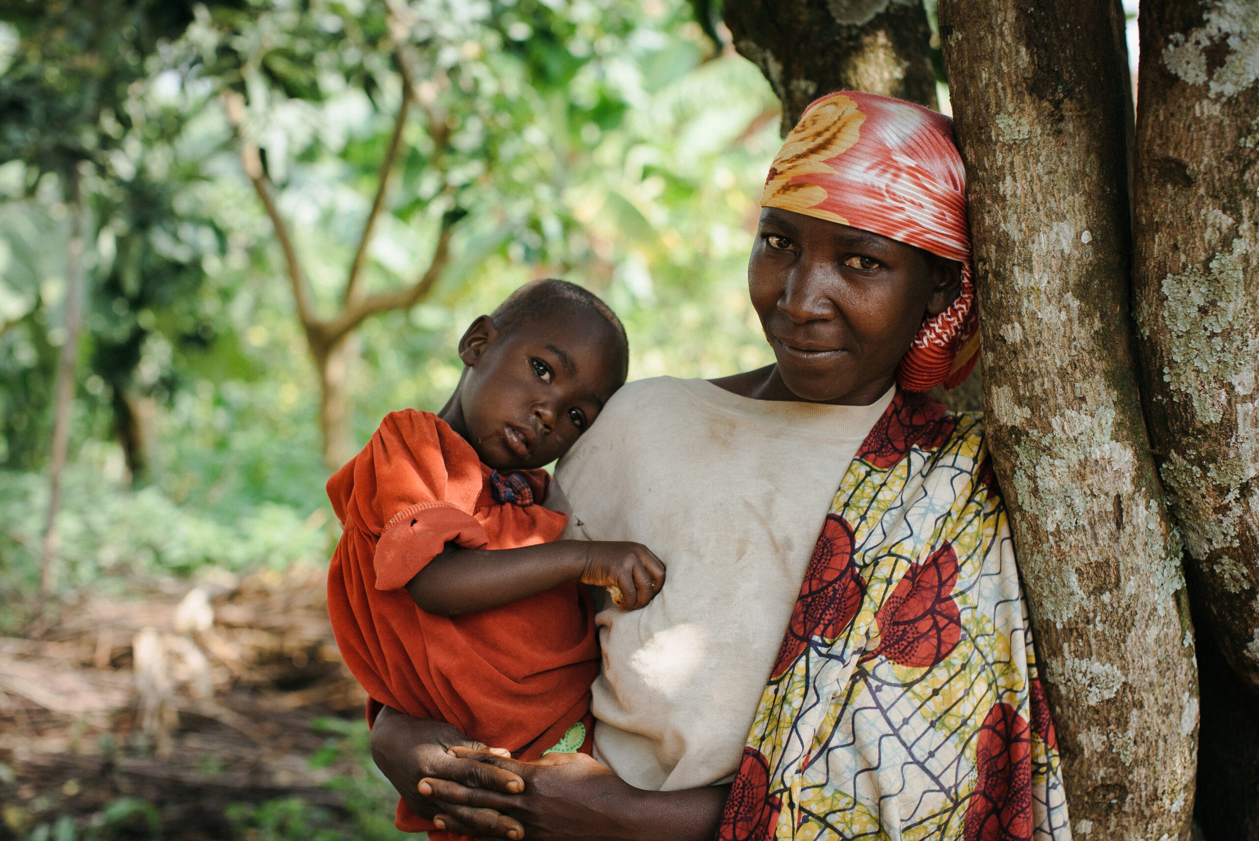 A woman wearing a colorful headscarf and patterned wrap holds a young child dressed in a red shirt. They stand next to a tree in a lush, green outdoor setting, with the woman leaning against the tree while the child rests against her shoulder.