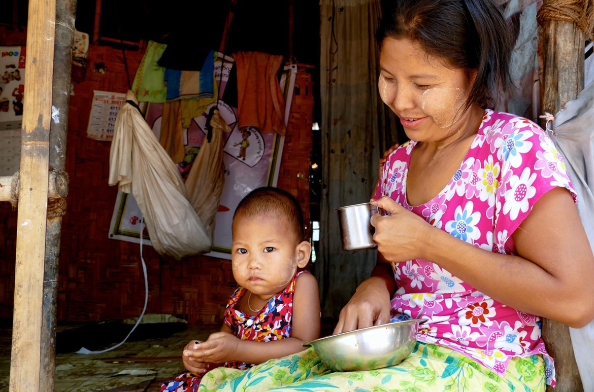 A woman and a child sit at the entrance of a woven bamboo house. The woman, in a colorful floral dress, smiles while holding a metal cup and bowl. The child, also in a floral dress, looks towards the camera. Clothes hang in the background.