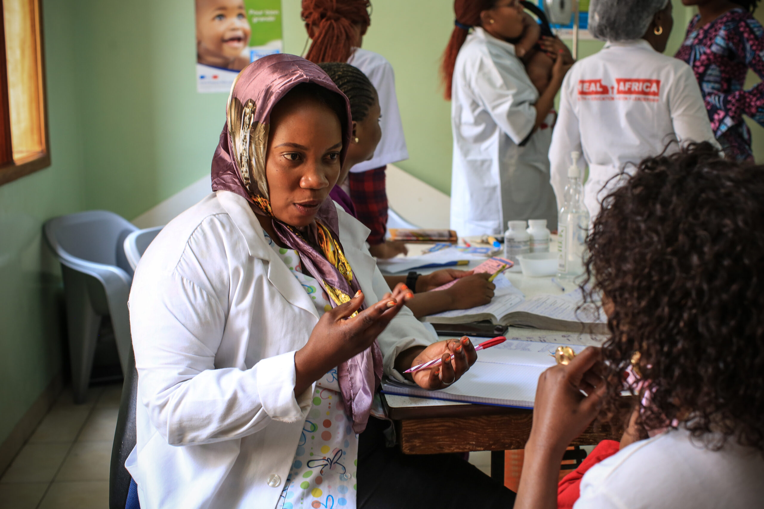 A female healthcare worker in a white coat and headscarf is talking to a patient while holding medication. In the background, other healthcare workers are assisting patients at a clinic. The setting appears busy and dedicated to patient care.