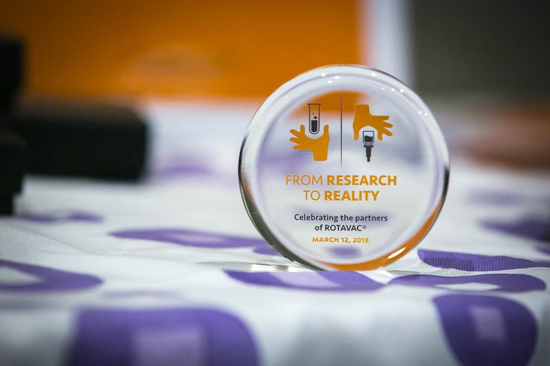 A circular glass award stands on a surface, featuring the text "FROM RESEARCH TO REALITY, Celebrating the partners of ROTAVAC®, MARCH 12, 2018" with icons of a test tube and a vaccine syringe flanked by illustrative hands.