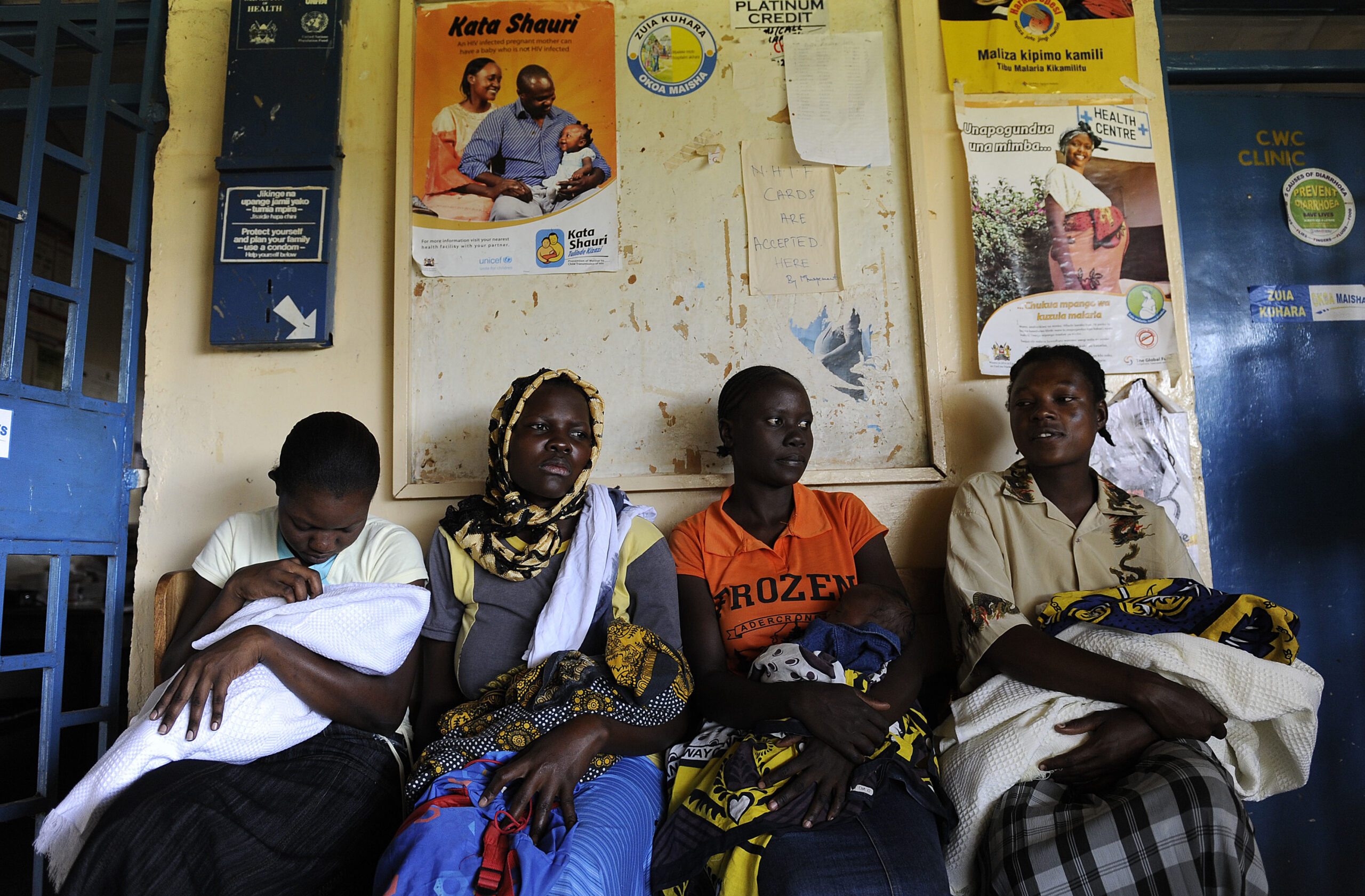 Four women sit on a bench, each holding an infant wrapped in colorful cloth. Behind them are various posters on a wall. The room appears to be a clinic waiting area.