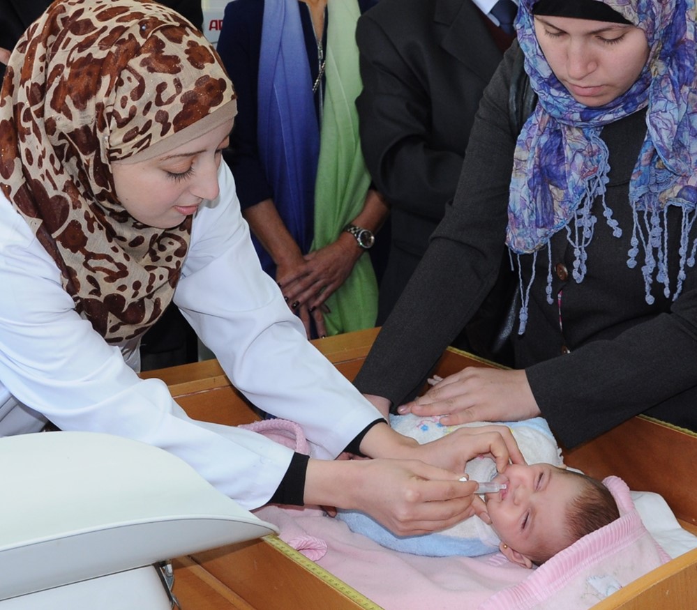 A healthcare professional in a white coat administers oral medication to a baby lying on a pink blanket, while a woman assists by holding the baby. Both women are wearing headscarves. The setting appears to be a medical facility with other people present in the background.