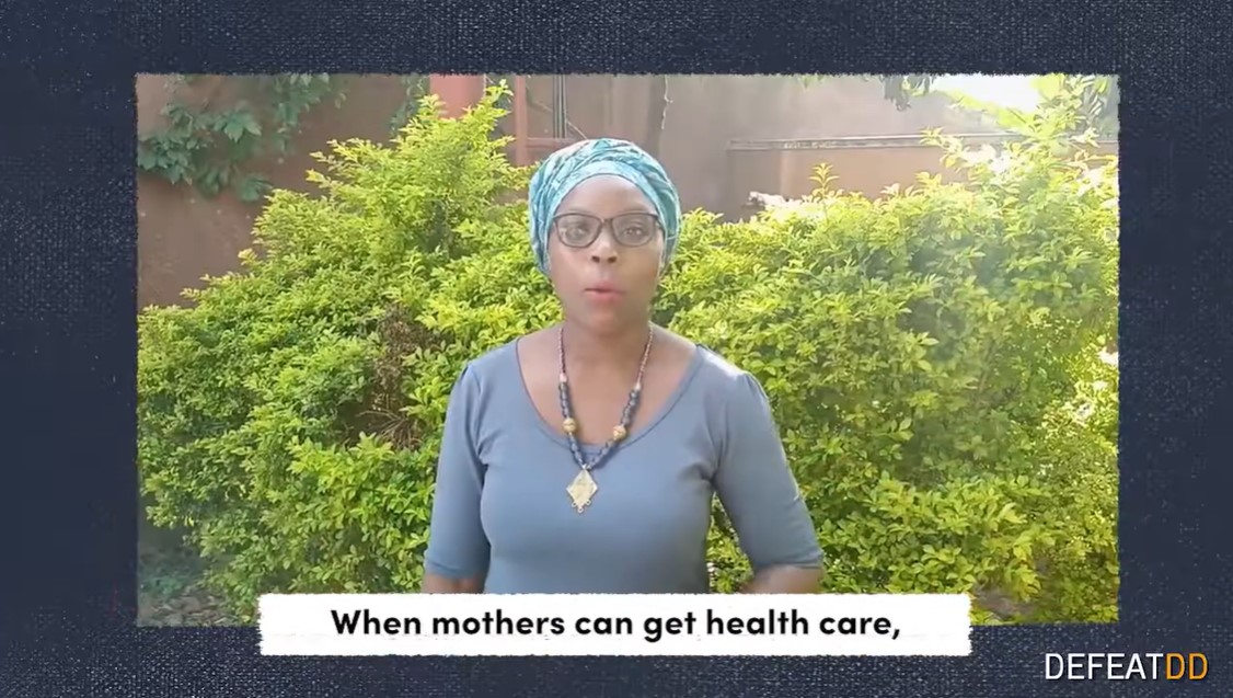 A woman wearing glasses and a blue headscarf is standing in front of a lush green bush. Text at the bottom of the image reads, "When mothers can get health care," with the logo "DEFEATDD" in the corner.