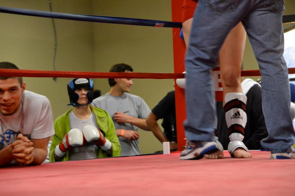 A person in a green jacket and boxing gear sits in a boxing ring corner while a kneeling individual in white leans on the ropes. Another person in jeans stands and a few others observe from outside the ring. The setting appears to be an indoor gym.