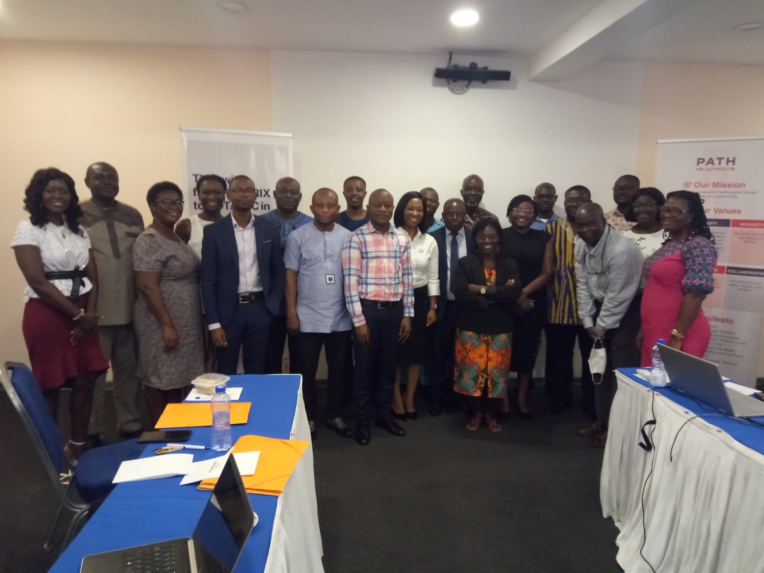 A diverse group of men and women, dressed in professional attire, stand together in a meeting room, smiling at the camera. There are banners in the background with the text "PATH" and some tables with laptops and documents in the foreground.