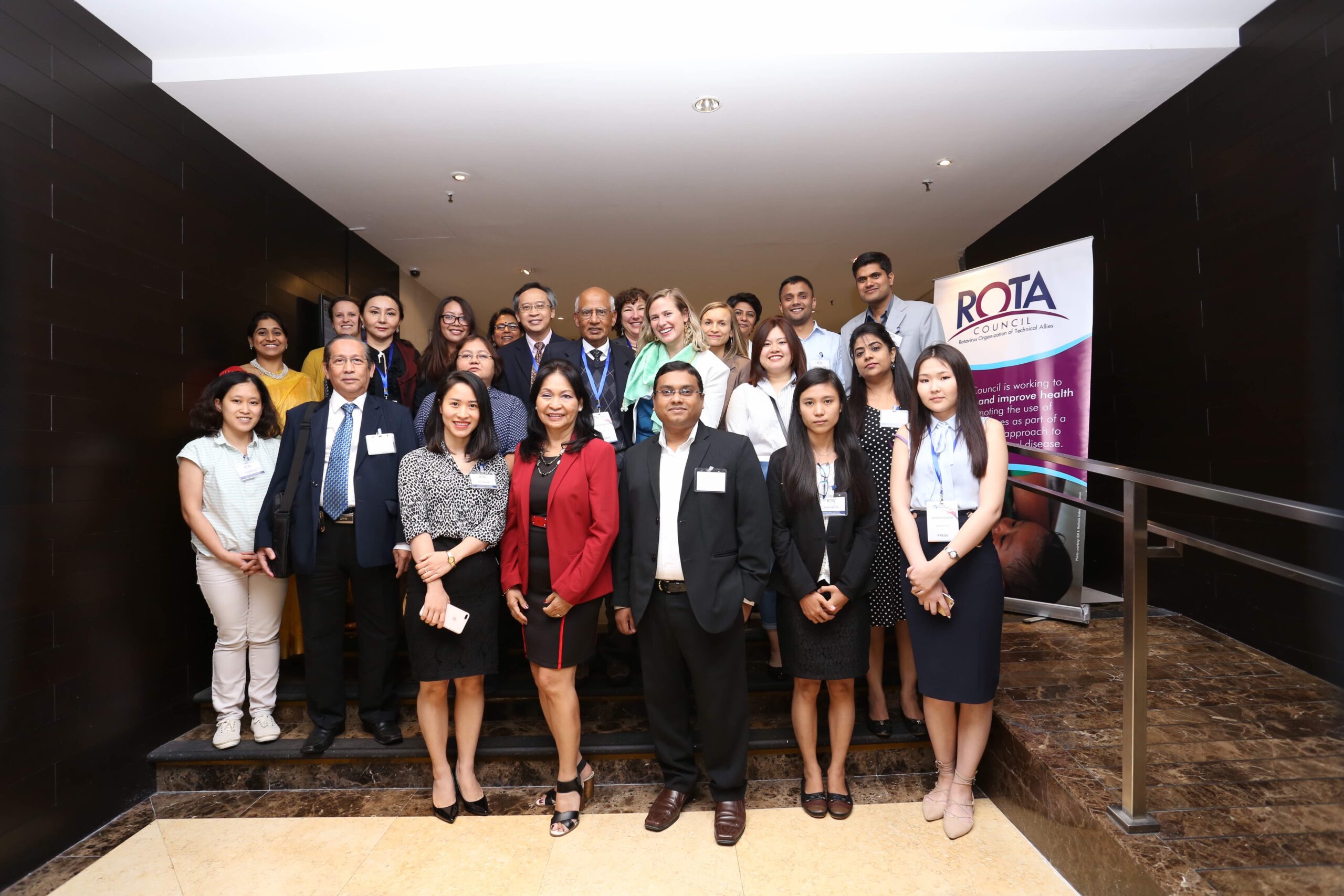 A group of diverse people is standing on stairs indoors, smiling for a photo. A sign with "ROTA" is visible on the right. They appear to be attending a formal or corporate event.