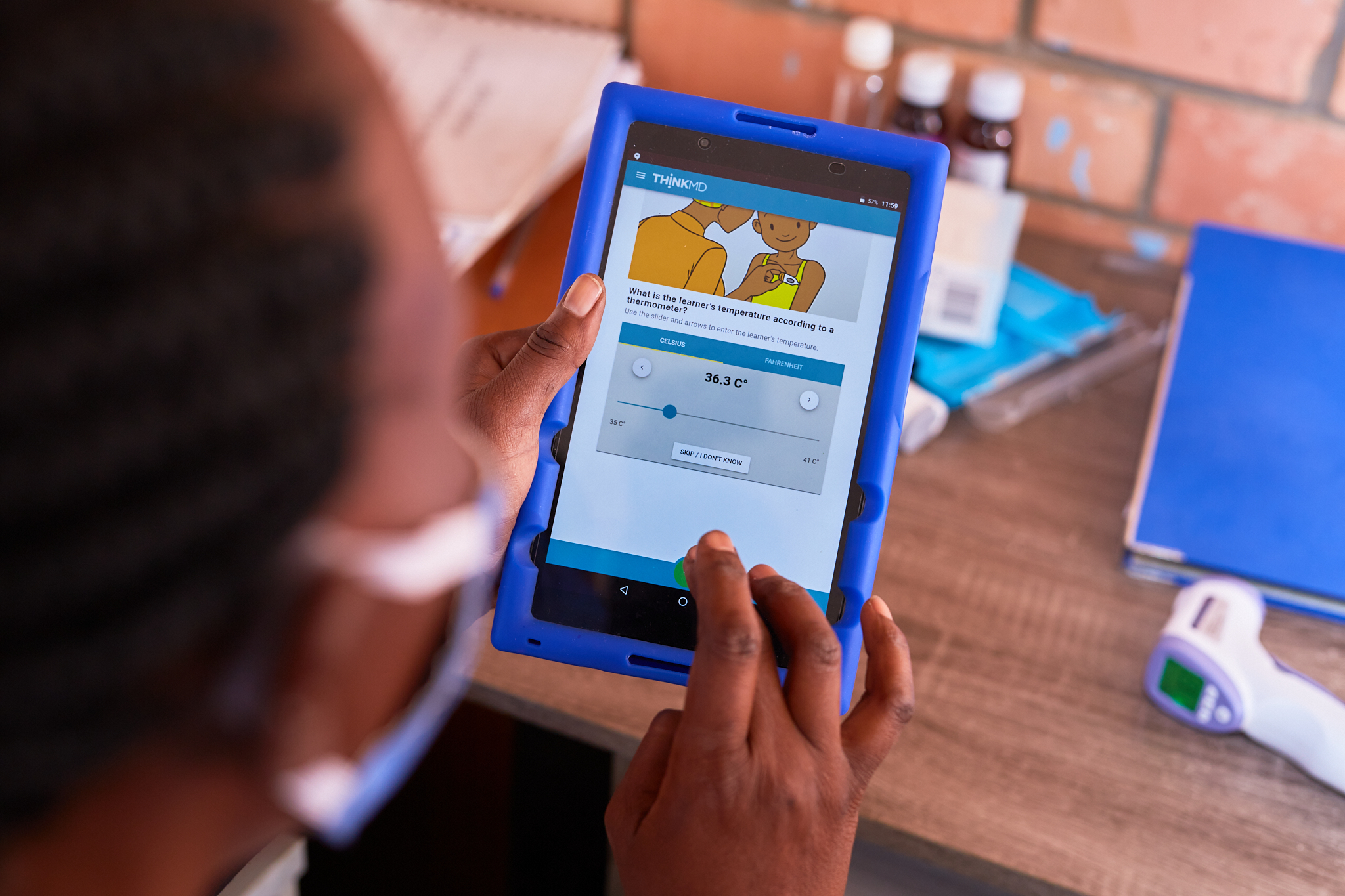 A person using a blue tablet to measure temperature is seen under a shaded element. The screen displays a temperature reading of 36.3°C. The background includes a desk with a thermometer, medical supplies, and a brick wall.