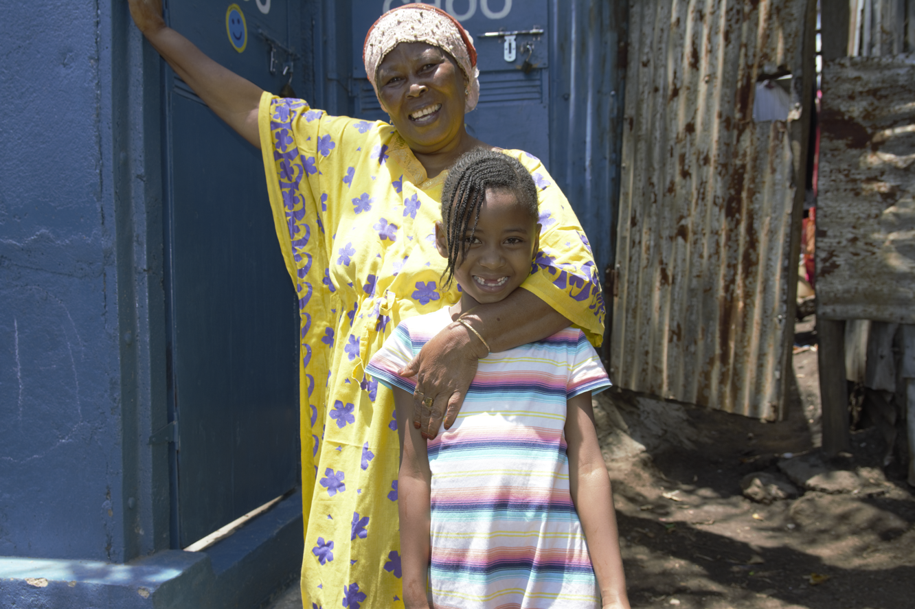 A woman in a yellow floral dress and headscarf stands with her arm around a smiling girl in a striped shirt. They are standing outdoors in front of a blue door and a corrugated metal wall. The background is sunlit and rustic.