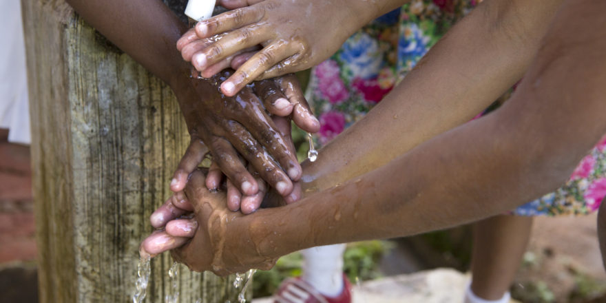 Children's hands of different skin tones being washed together under an outdoor faucet. The background shows part of a wooden post and someone's floral print clothing. Water flows over their hands, indicating a shared and communal activity.