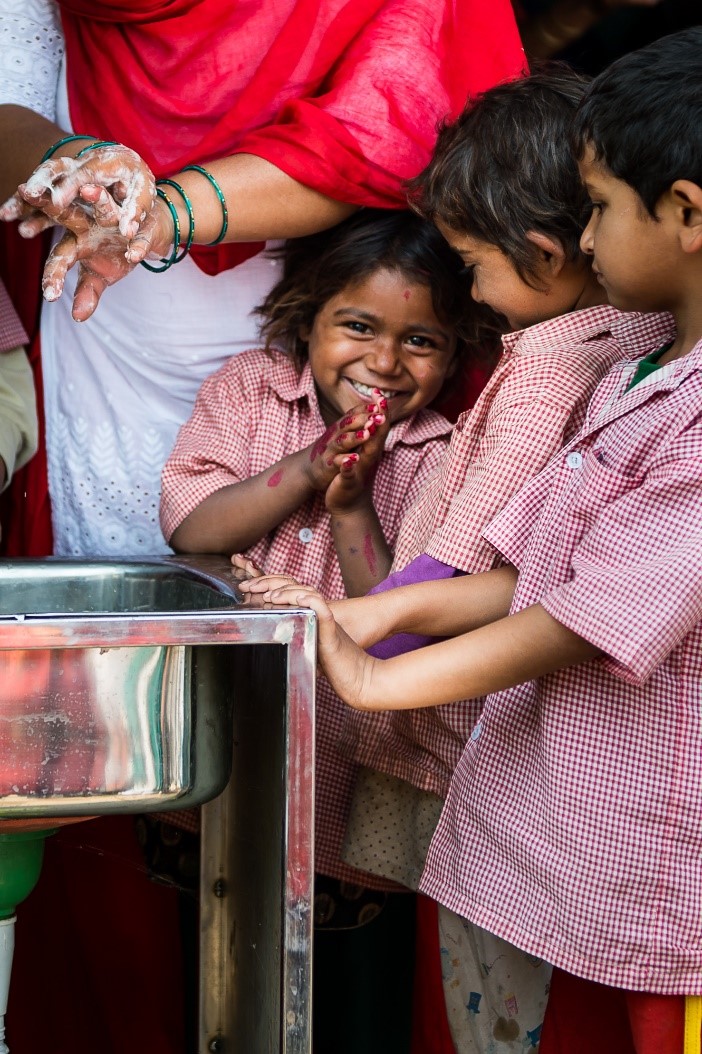 Three children in checkered uniforms stand at a metal sink. The central child, smiling, clasps her hands with soapy water, while the child on the right looks on. An adult in a red shawl assists them, reinforcing the importance of handwashing.