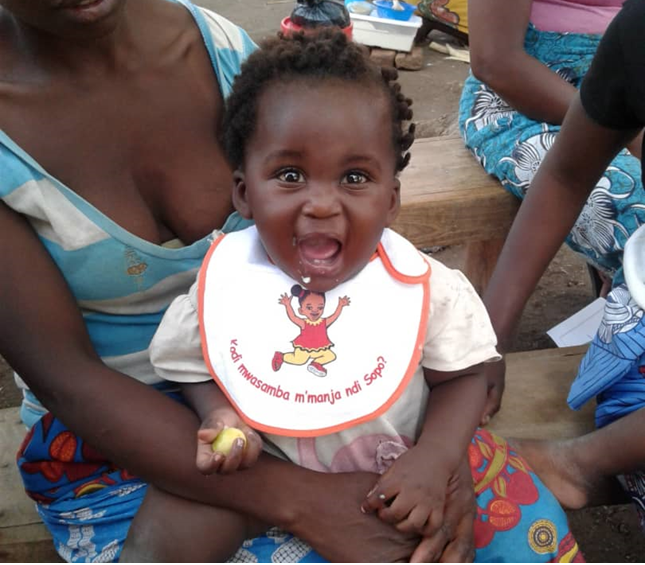 A cheerful baby with curly hair sits on an adult's lap, wearing a white bib with a cartoon image and red text. The child is holding a small object, possibly a fruit. There are two adults, partially visible, sitting nearby on a wooden bench.