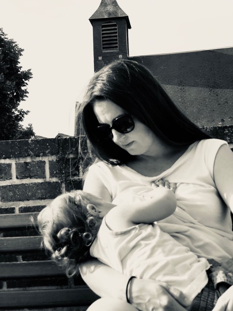 A woman with long dark hair and sunglasses sits on a bench breastfeeding a young child. There is a brick wall behind them, with a building and a steeple in the background. The image is in black and white.