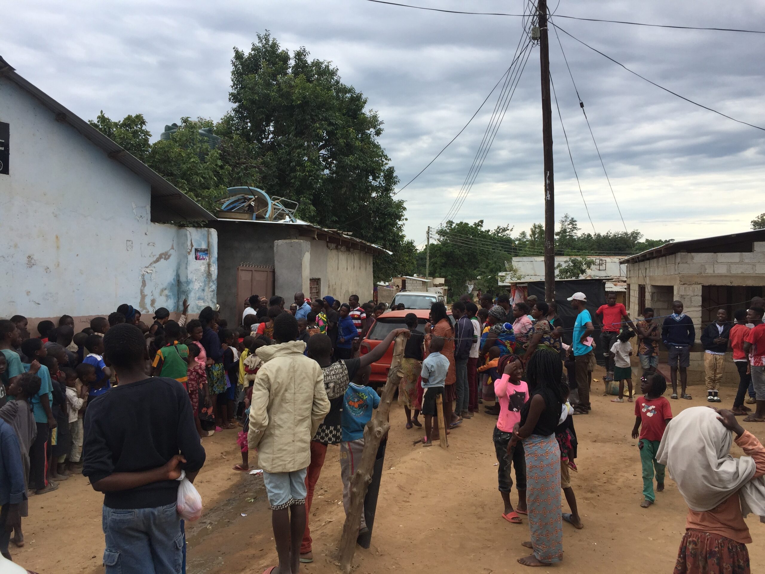 A large group of people, including many children, gather in a residential area with modest houses and an unpaved road. A cloudy sky looms overhead, and several people appear to be engaged in conversation or waiting for something. Power lines are visible.