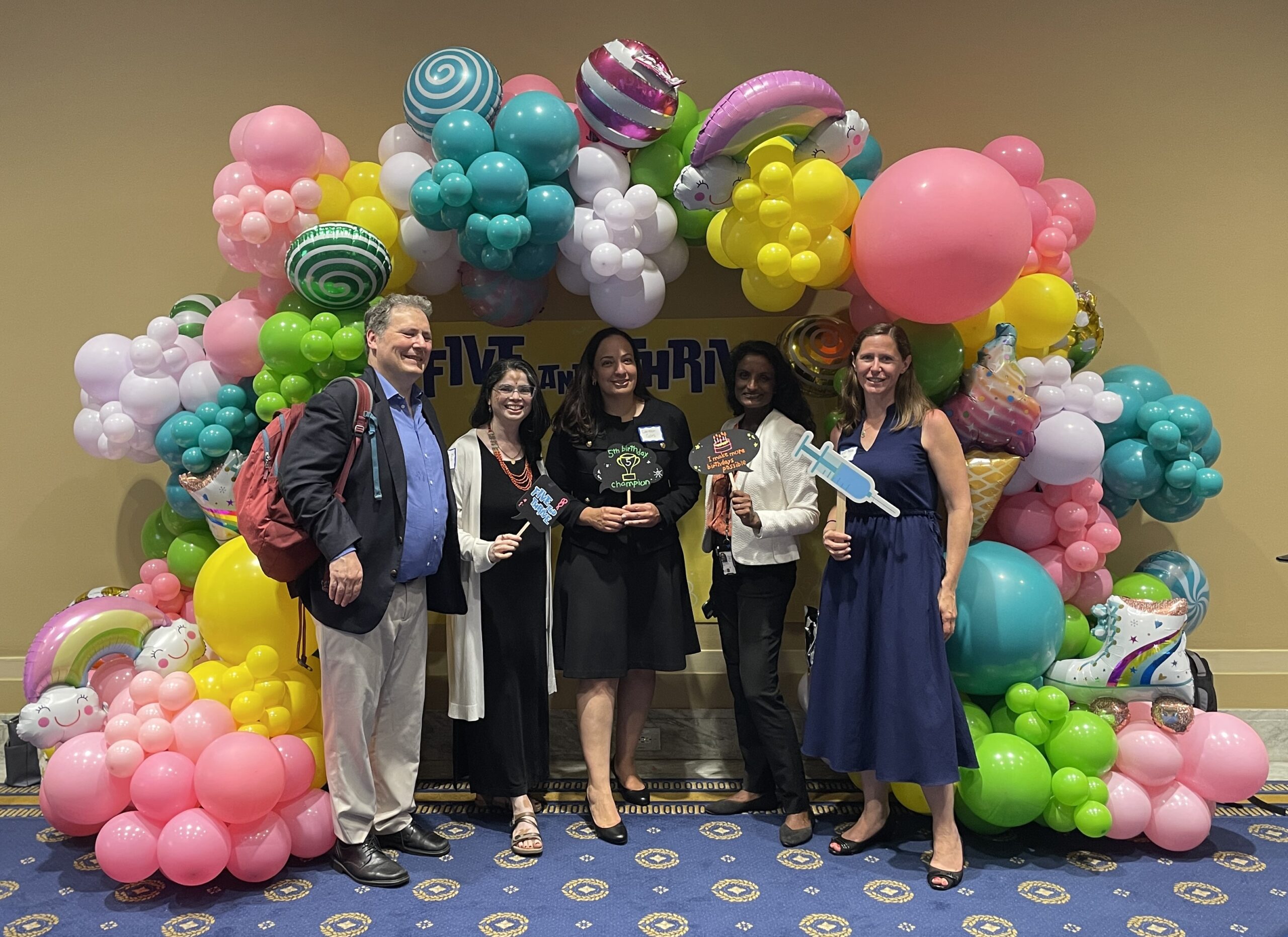 A group of five adults stands in front of a colorful balloon arch at a festive event. The arch features balloons in various shapes and colors, including flowers, stars, and swirls. All are smiling and holding props, and one person is wearing a backpack.