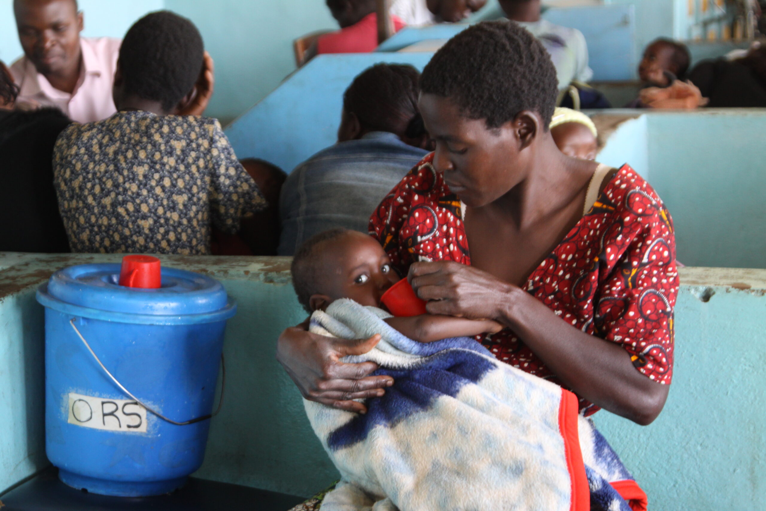 A woman in a red patterned dress sits holding a young child wrapped in a blanket, feeding them from a red cup. They are in a crowded room with blue walls, and a blue container labeled "ORS" is visible beside them. Several other people are seen in the background.