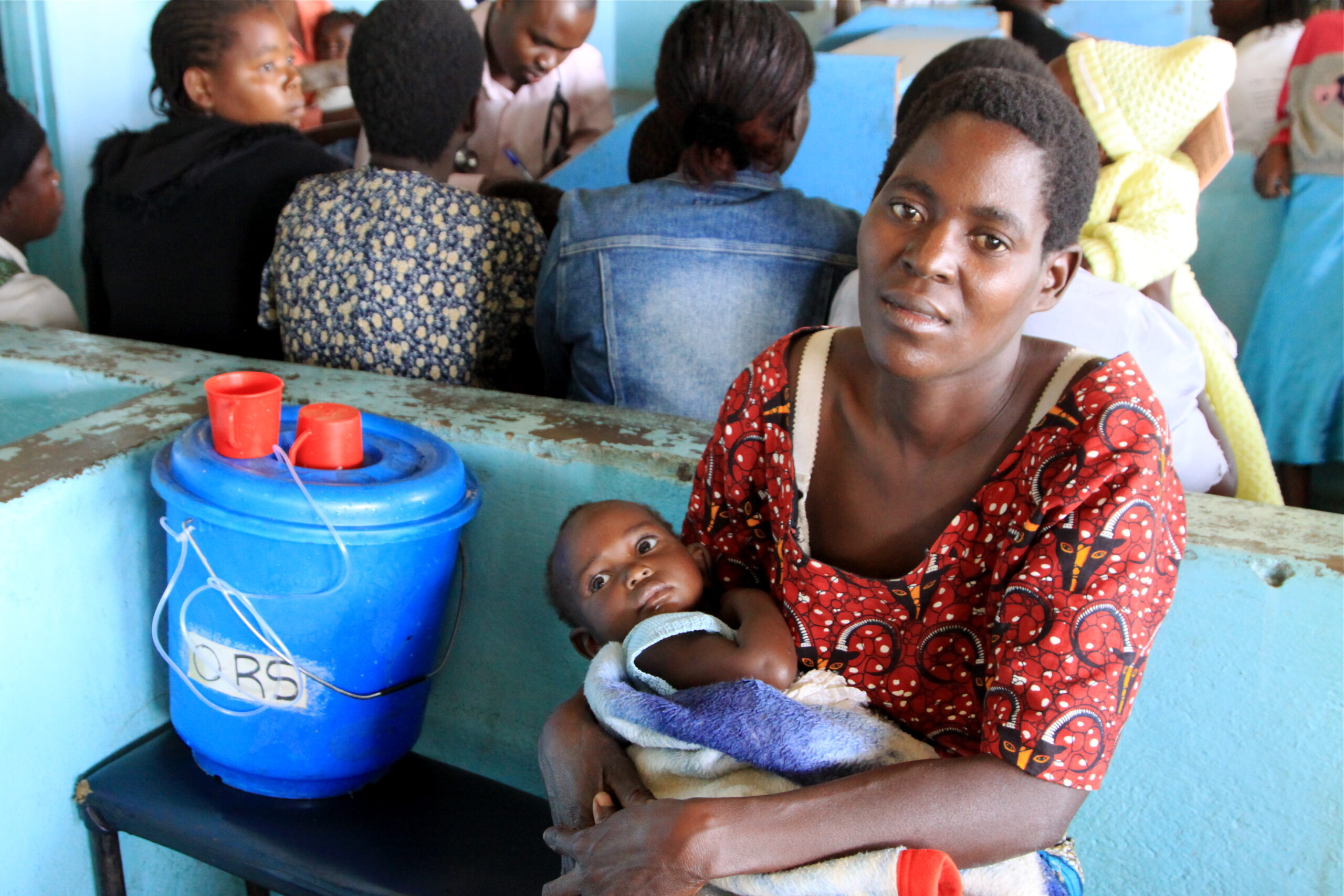 A woman wearing a red patterned dress holds a baby wrapped in a blanket while sitting on a bench. In front of her is a blue container labeled "ORS." Several people sit and talk in the background. The setting appears to be a healthcare facility.