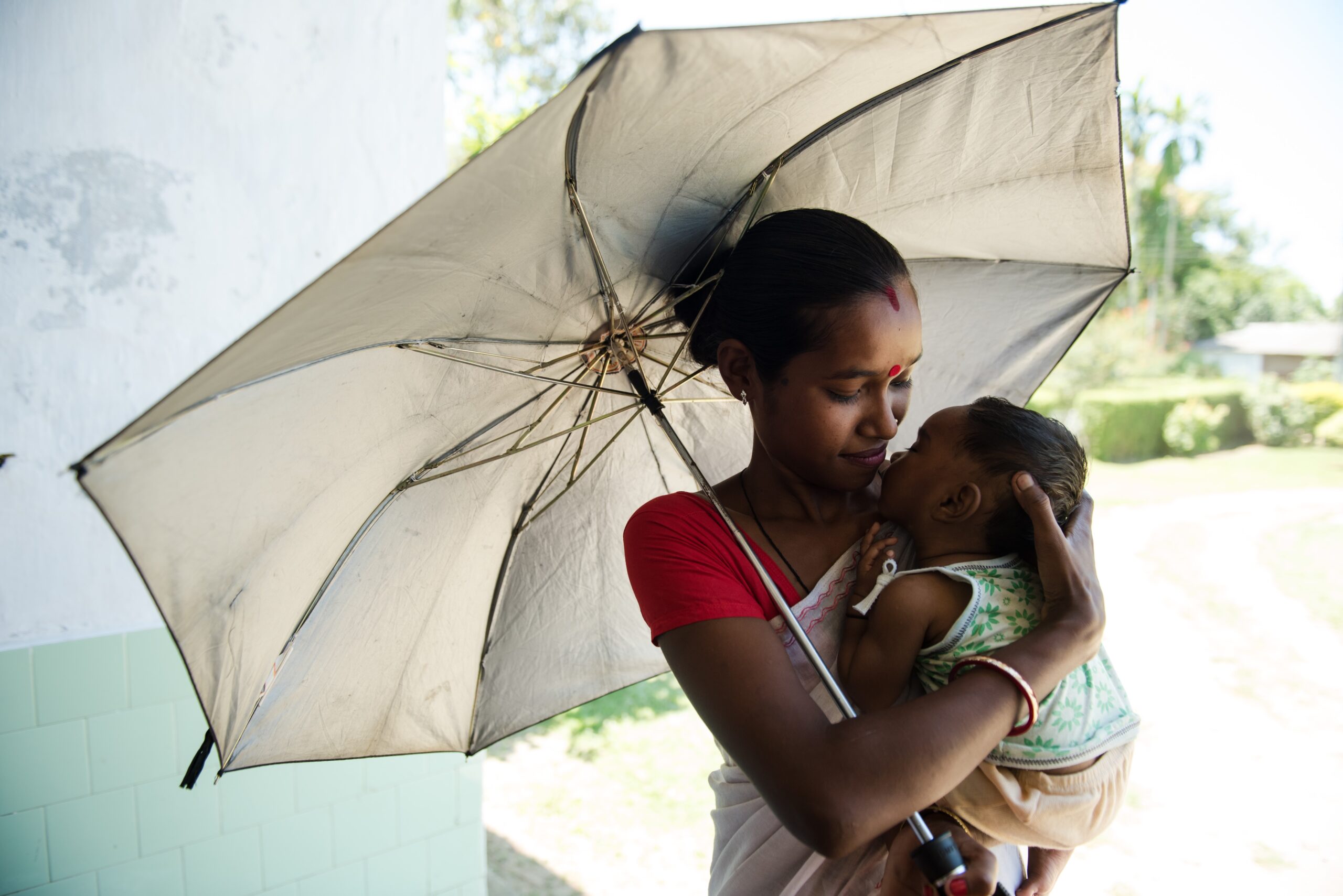 A woman gently holds a baby under a large umbrella. She is wearing a traditional sari and jewelry. They are standing outside, with greenery in the background. The scene is filled with soft, natural light.