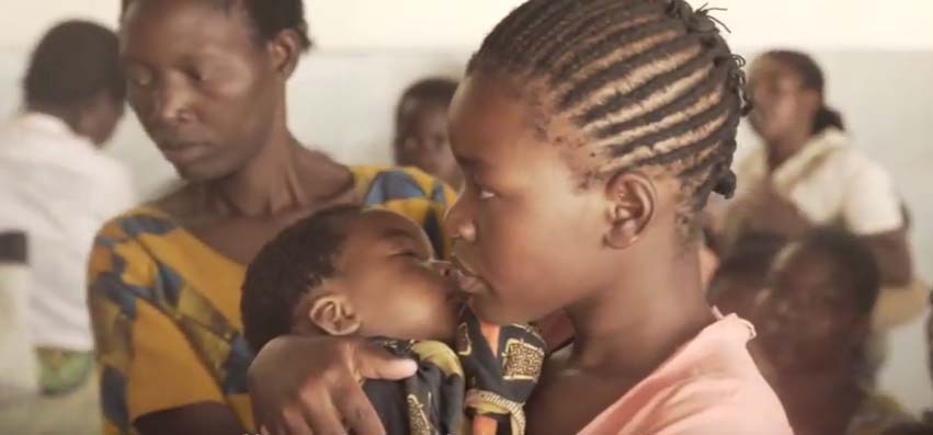 A young girl with neatly braided hair holds and comforts a baby in her arms. They are indoors among other people, including a woman in the background who is wearing a patterned dress and looking downward. The setting appears to be a communal or medical facility.