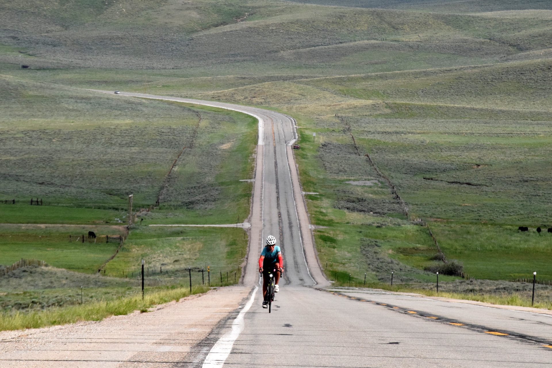 A lone cyclist rides up a long, straight road that stretches into the distance, surrounded by vast, rolling green hills under an overcast sky. The road appears empty and tranquil, emphasizing the solitary nature of the cyclist's journey.