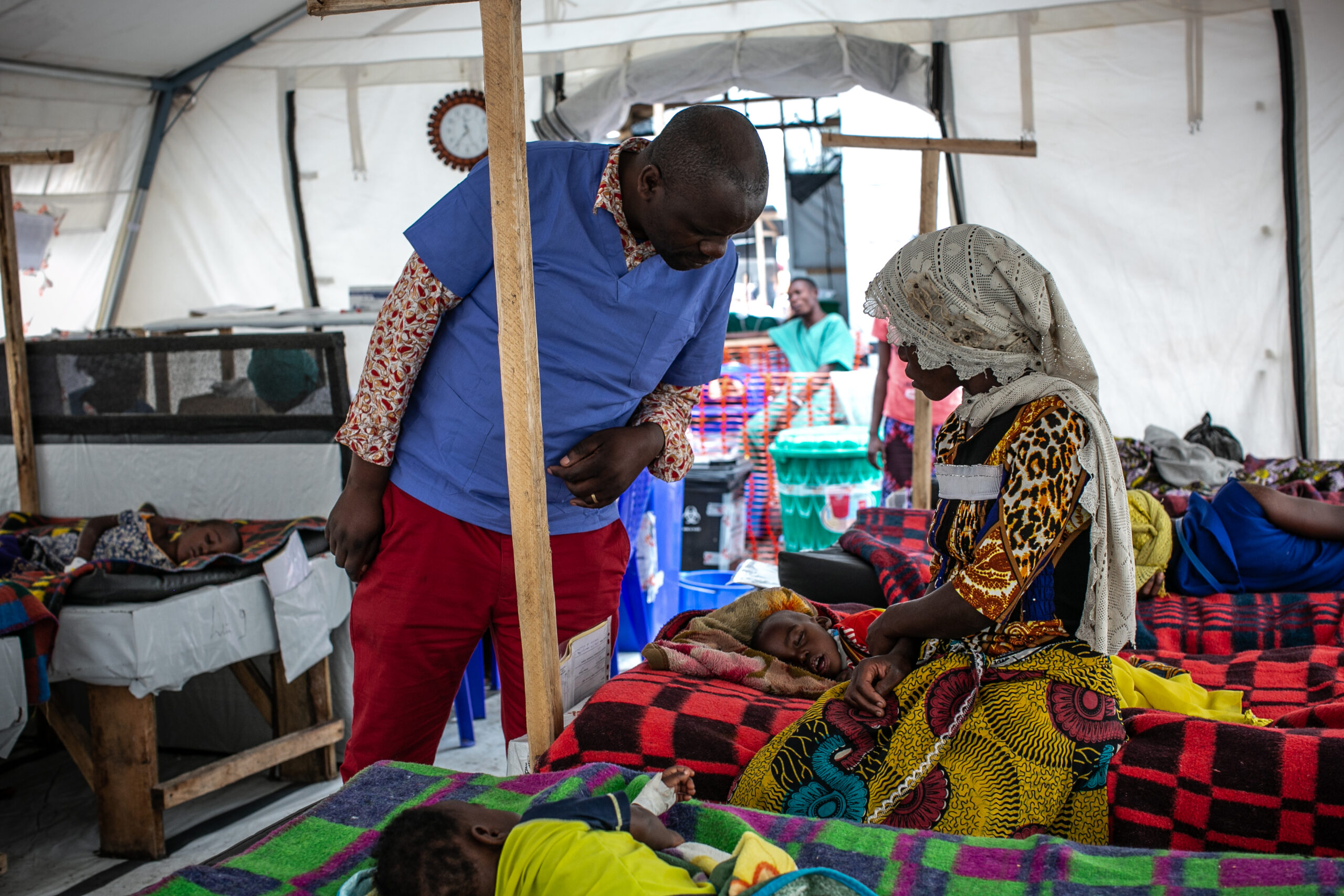 A healthcare worker in blue scrubs and red pants talks to a woman in colorful attire and a headscarf, who is seated next to a child lying on a bed in a medical tent. Other patients are seen resting on beds in the background.