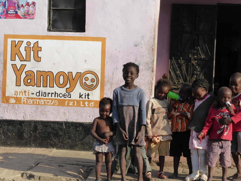 Seven children stand in front of a building painted with a sign for "Kit Yamoyo - anti-diarrhoea kit". The children are dressed in casual clothes, some holding toys or bottles, and they appear to be posing for the photo. The building has a pale wall and a metal door.