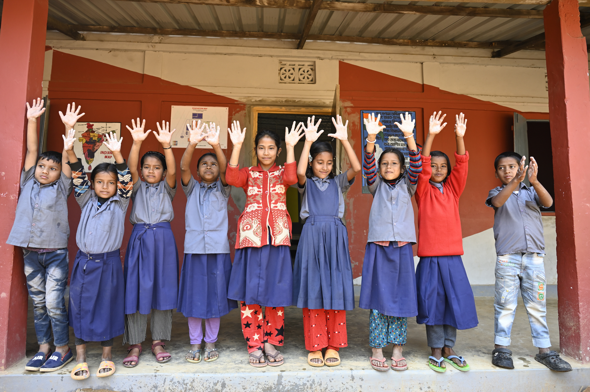 A group of children stands in a row in front of a building with their arms raised high, smiling. The children are dressed in various outfits, including uniforms and casual clothes. The backdrop shows a wall with posters and red and white paint.