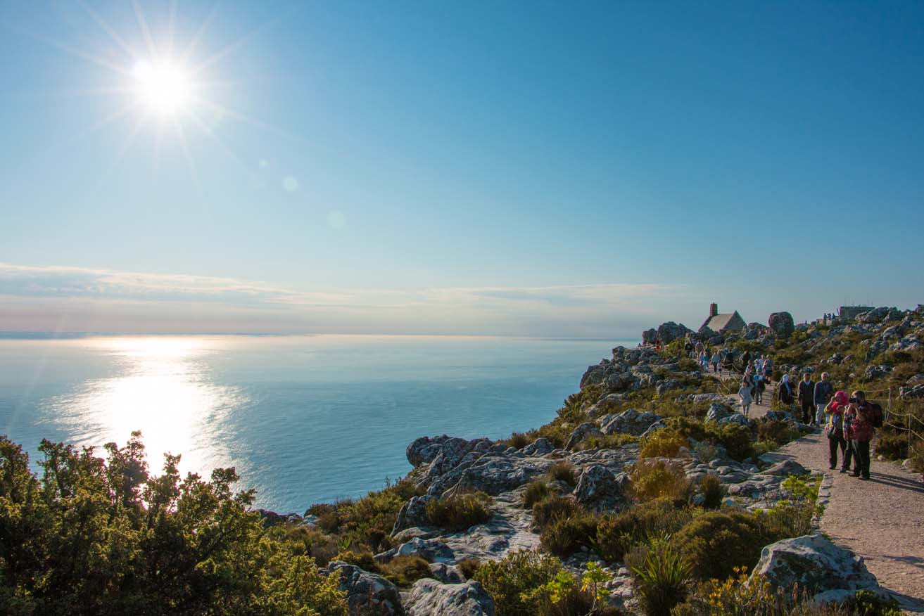 A scenic view of a rocky coastline bathed in sunlight, with a clear blue sky and the sun shining brightly overhead. People are walking along a path, enjoying the view of the calm ocean and the rugged landscape. Shrubs and rocks are scattered along the cliffside.